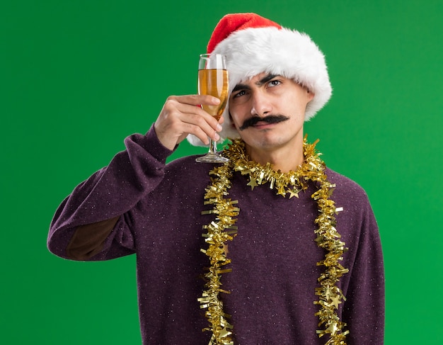 Giovane uomo baffuto che indossa il cappello di Babbo Natale di Natale con orpelli intorno al collo tenendo il bicchiere di champagne guardando perplesso