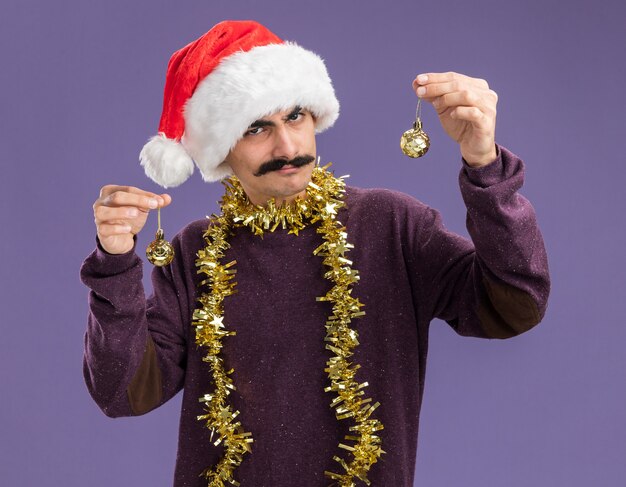 Giovane uomo baffuto indossando natale santa hat con orpelli intorno al collo tenendo le palle di natale guardando la fotocamera con espressione scettica in piedi su sfondo viola