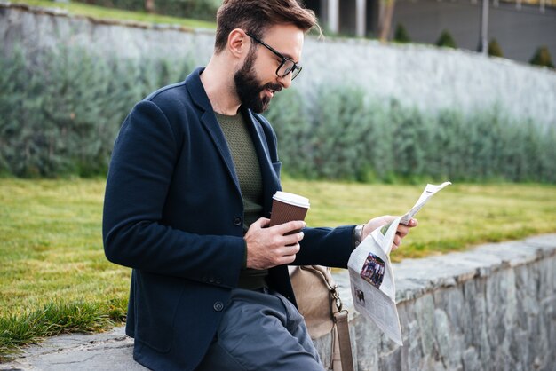 Giovane uomo barbuto bello che si siede all'aperto leggendo giornale