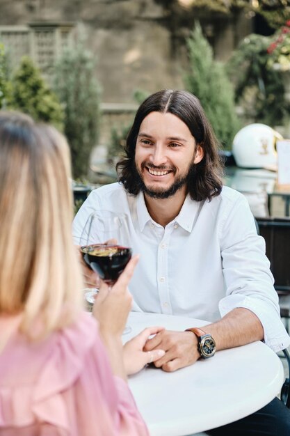 Giovane uomo barbuto bruna attraente in camicia con bicchiere di vino che guarda con gioia la ragazza in un appuntamento romantico nella caffetteria all'aperto