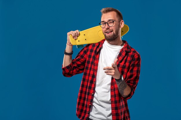 Giovane uomo barbuto carismatico allegro vestito con camicia a scacchi, maglietta bianca e occhiali, con skateboard giallo nelle sue mani. Studio girato con sfondo blu. Fricchettone