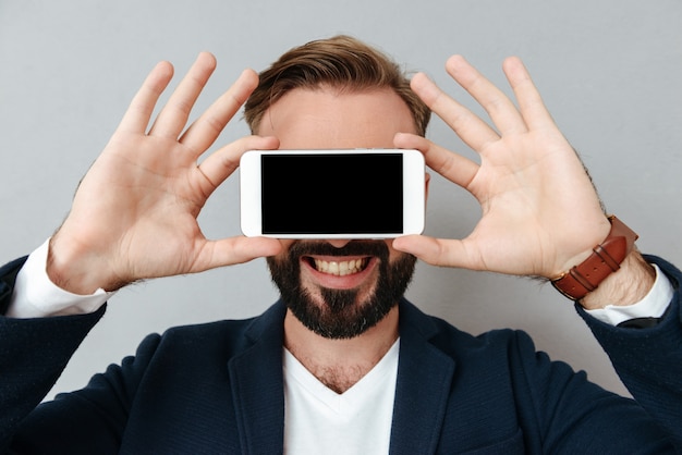 Giovane uomo barbuto che copre il viso con lo smartphone con schermo vuoto