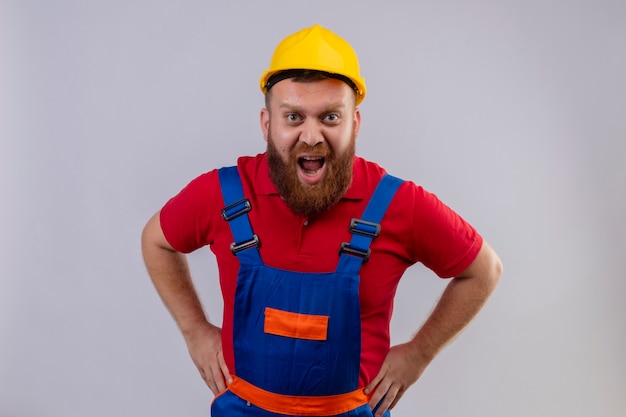 Giovane uomo barbuto costruttore in uniforme da costruzione e casco di sicurezza che grida con espressione aggressiva