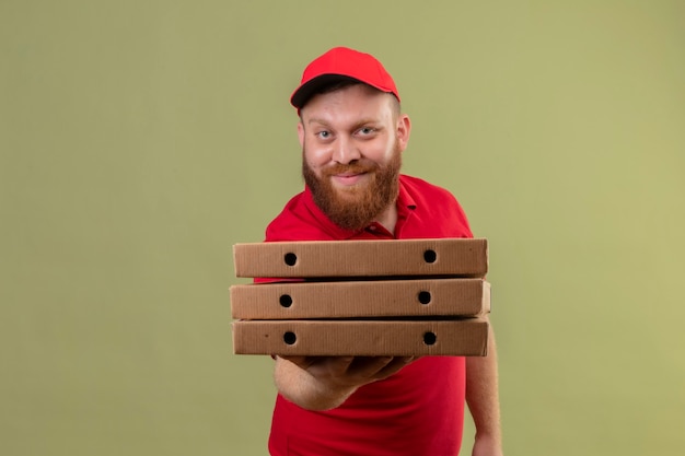 Giovane uomo barbuto di consegna in uniforme rossa e cappuccio che tiene pila di scatole per pizza sorridente amichevole