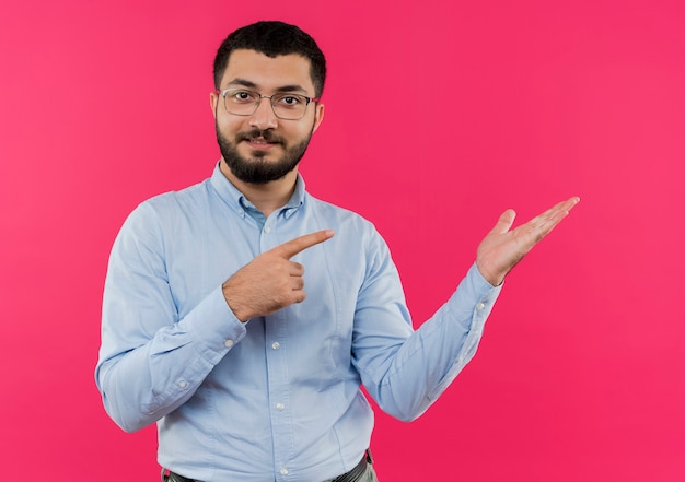 Giovane uomo barbuto in bicchieri e camicia blu che presenta qualcosa con il braccio rivolto con il dito indice al lato sorridente fiducioso