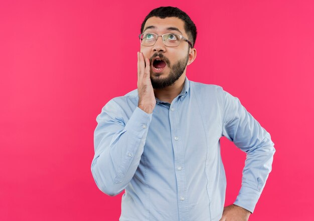 Giovane uomo barbuto in bicchieri e maglietta blu alzando lo sguardo di essere scioccato