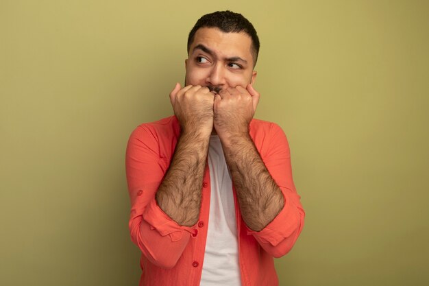 Giovane uomo barbuto in camicia arancione che osserva da parte unghie mordaci stressate e nervose in piedi sopra la parete leggera