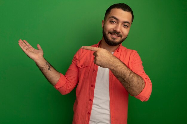 Giovane uomo barbuto in camicia arancione che presenta qualcosa con il braccio rivolto con il dito indice a lato sorridente fiducioso in piedi oltre la parete verde