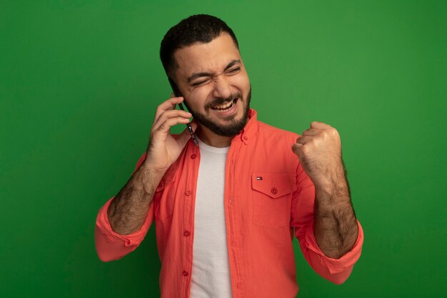 Giovane uomo barbuto in camicia arancione che stringe il pugno felice ed eccitato mentre parla al telefono cellulare in piedi sopra la parete verde