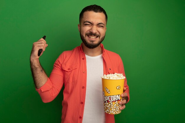 Giovane uomo barbuto in camicia arancione che tiene secchio con popcorn e tv a distanza felice ed emozionato alzando la mano in piedi sopra la parete verde