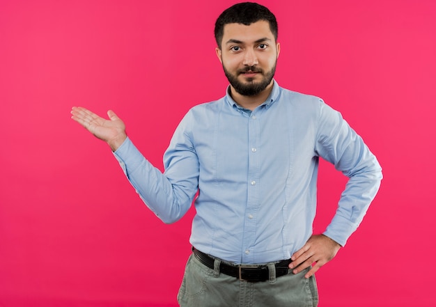 Giovane uomo barbuto in camicia blu che presenta qualcosa con il braccio della sua mano che sembra fiducioso