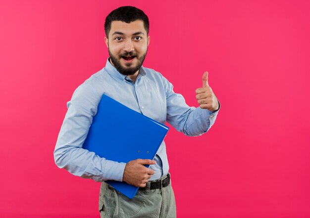 Giovane uomo barbuto in camicia blu che tiene cartella sorridente che mostra i pollici in su