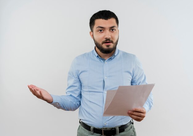 Giovane uomo barbuto in camicia blu che tiene pagine vuote guardando davanti con il braccio fuori essendo scontento in piedi sul muro bianco