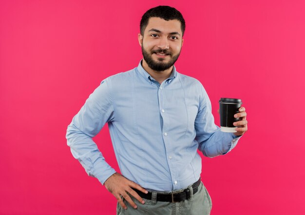 Giovane uomo barbuto in camicia blu che tiene tazza di caffè sorridente con la faccia felice