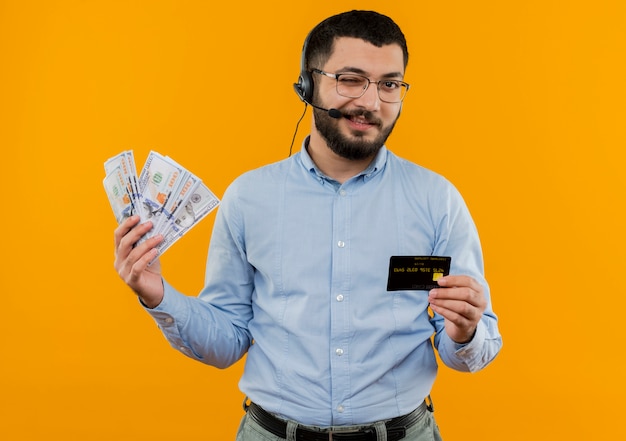 Giovane uomo barbuto in camicia blu con cuffie con microfono che tiene contanti che mostra carta di credito sorridente e ammiccante