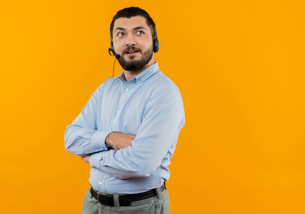 Giovane uomo barbuto in camicia blu con le cuffie con microfono che osserva da parte con espressione pensierosa con le braccia incrociate