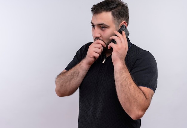 Giovane uomo barbuto in camicia nera che racconta un segreto mentre parla al telefono cellulare