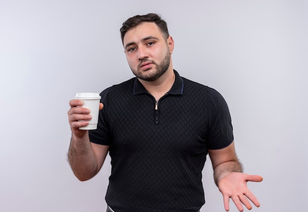 Giovane uomo barbuto in camicia nera che tiene tazza di caffè che sembra confuso