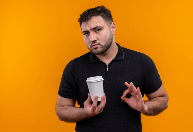 Giovane uomo barbuto in camicia nera che tiene tazza di caffè facendo segno ok guardando fiducioso