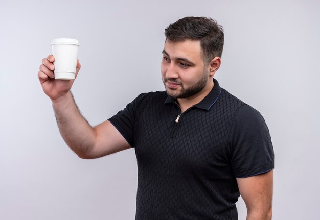 Giovane uomo barbuto in camicia nera tenendo la tazza di caffè alzando la mano sorridente fiducioso