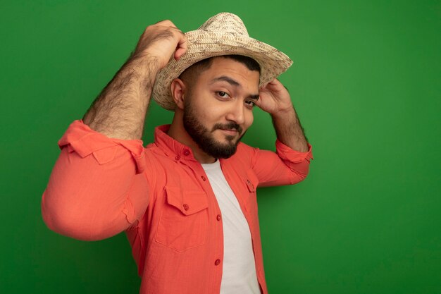 Giovane uomo barbuto in maglietta arancione e cappello estivo guardando sorridente fiducioso che fissa il suo cappello in piedi sopra la parete verde