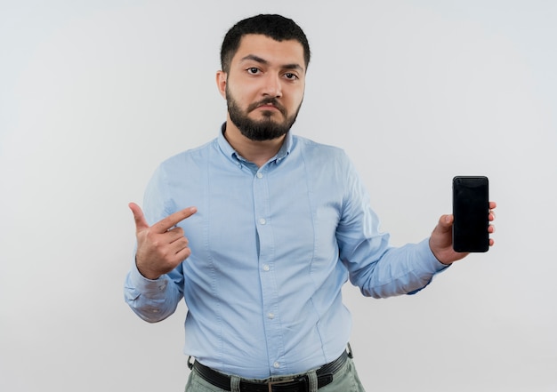 Giovane uomo barbuto in maglietta blu che tiene smartphone che punta con il dito indice a essere confuso