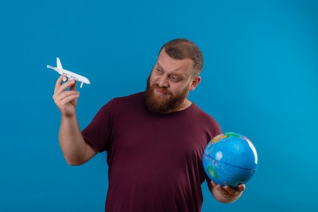 Giovane uomo barbuto in maglietta marrone che tiene globo e aeroplano giocattolo guardando il giocattolo con l'espressione scettica sul viso