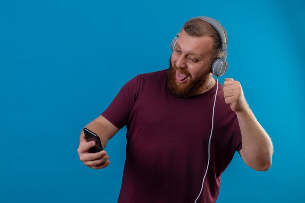Giovane uomo barbuto in maglietta marrone con le cuffie prendendo selfie utilizzando il suo smartphone facendo smorfia che attacca fuori la lingua