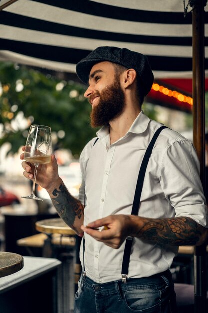 Giovane uomo barbuto in un caffè per strada con un bicchiere di vino. Ragazzo romantico con un berretto da camicia bianca e bretelle in città. Peaky Blinders. vecchia moda retrò.