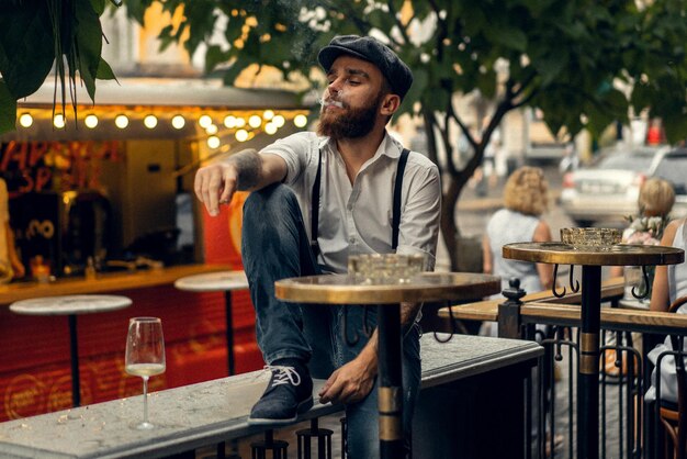 Giovane uomo barbuto in un caffè per strada con un bicchiere di vino. Ragazzo romantico con un berretto da camicia bianca e bretelle in città. Peaky Blinders. vecchia moda retrò.