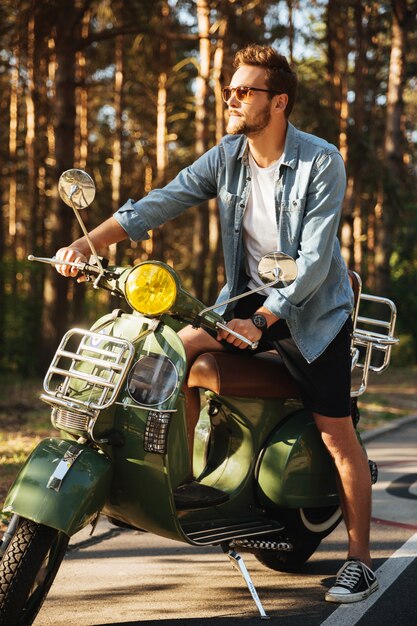 Giovane uomo barbuto serio bello che si siede sul motorino