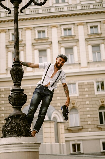 Giovane uomo barbuto tatuato. Un ragazzo romantico in camicia bianca, berretto e bretelle cammina per la città. ruota attorno a un lampione. Peaky Blinders. vecchio stile, retrò.