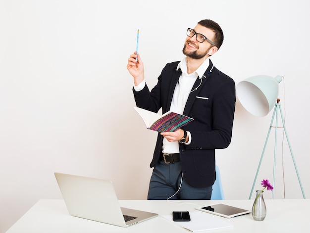 Giovane uomo bello alla moda hipster in giacca nera che lavora al tavolo dell'ufficio, stile aziendale, camicia bianca, isolato, laptop, avvio, posto di lavoro, pensiero, documenti