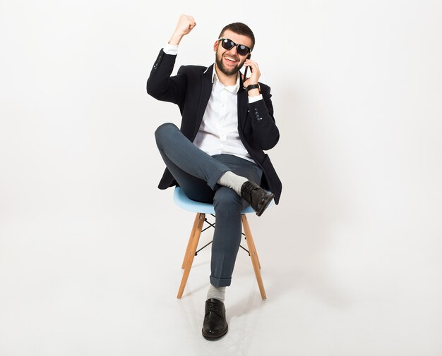 Giovane uomo bello alla moda hipster in giacca nera, stile di affari, camicia bianca, isolato, seduto rilassato sulla sedia da ufficio, parlando sullo smartphone, sorridente, felice, positivo, occhiali da sole