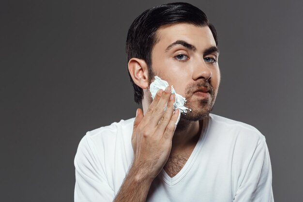 Giovane uomo bello che applica la crema per la rasatura sul viso su grigio.
