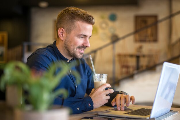 Giovane uomo bello che beve caffè mentre si utilizza il computer portatile nella caffetteria