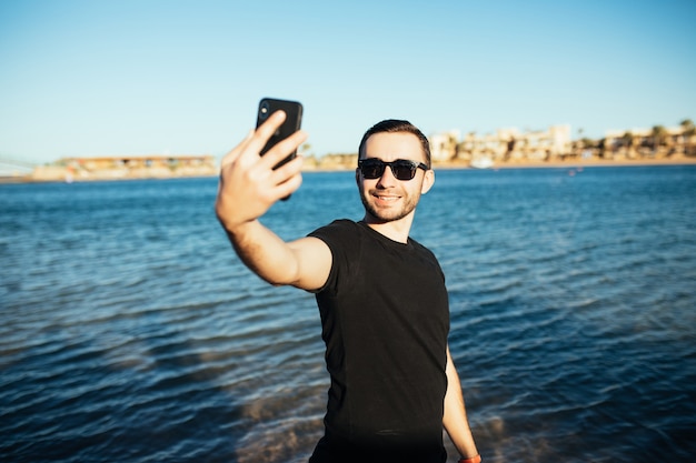 Giovane uomo bello che fa un autoritratto con lo smartphone sulla spiaggia