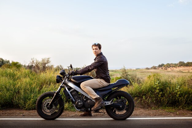 Giovane uomo bello che guida su una moto a strada di campagna.