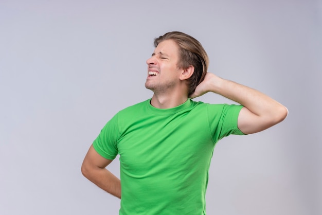 Giovane uomo bello che indossa la maglietta verde alla ricerca di malessere sensazione di dolore alla schiena in piedi sopra il muro bianco