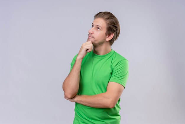 Giovane uomo bello che indossa la maglietta verde che osserva in su con la mano sul mento con l'espressione pensierosa sul fronte che sta sopra il muro bianco