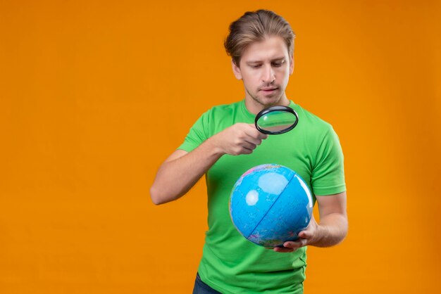 Giovane uomo bello che indossa la maglietta verde guardando il globo attraverso la lente di ingrandimento con la faccia seria in piedi sopra la parete arancione