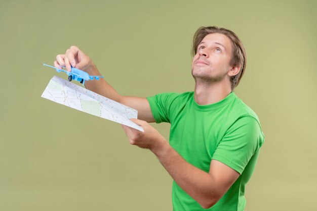 Giovane uomo bello che indossa t-shirt verde che tiene aeroplano giocattolo e mappa alzando lo sguardo sorridente in piedi sopra la parete verde