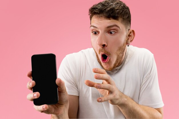 Giovane uomo bello che mostra lo schermo dello smartphone sopra lo spazio rosa con una faccia a sorpresa