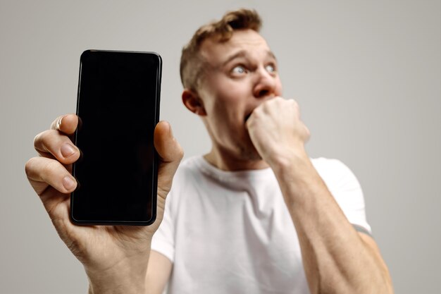 Giovane uomo bello che mostra lo schermo dello smartphone su grigio con una faccia a sorpresa.