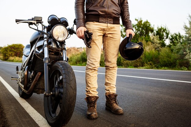 Giovane uomo bello che posa vicino alla sua motocicletta alla strada della campagna.