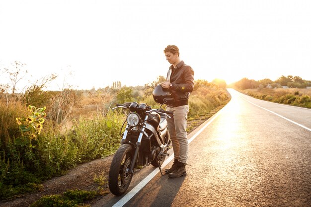 Giovane uomo bello che posa vicino alla sua motocicletta alla strada della campagna.