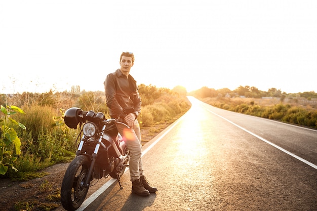 Giovane uomo bello che posa vicino alla sua motocicletta alla strada della campagna.