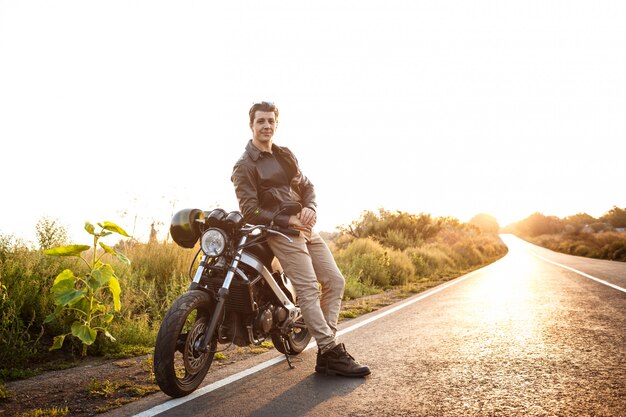 Giovane uomo bello che posa vicino alla sua motocicletta alla strada della campagna.