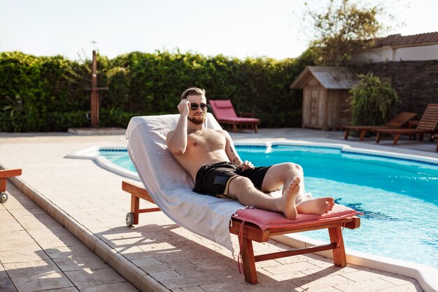 Giovane uomo bello che prende il sole, trovandosi sulle chaise vicino alla piscina