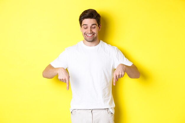 Giovane uomo bello che punta e guarda in basso, controllando l'offerta promozionale, in piedi su sfondo giallo.